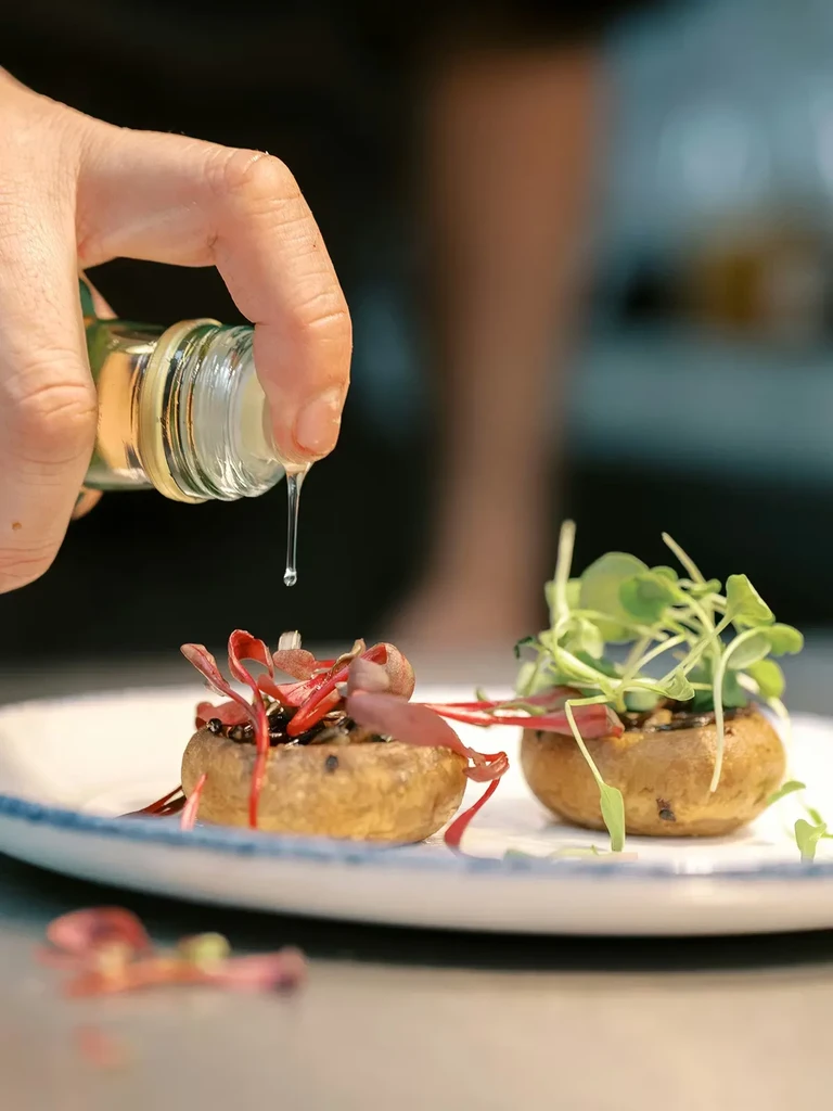 Chef's hand drizzles liquid over two appetizers with red and green microgreens.