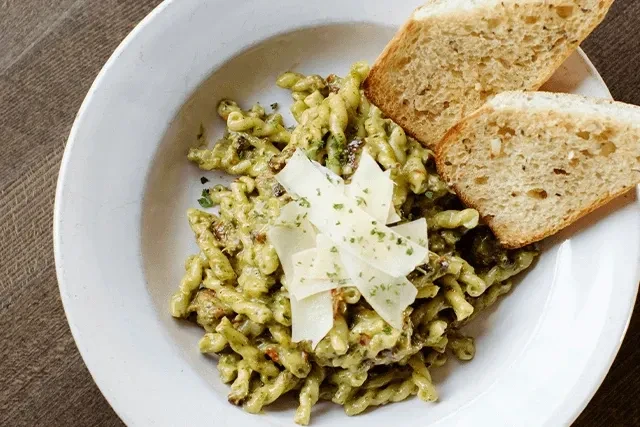 Overhead: Green pasta with cheese, herbs, and bread on a wooden table.