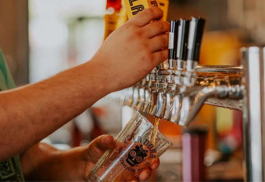 Hands pour amber beer from tap into skull-logo glass.