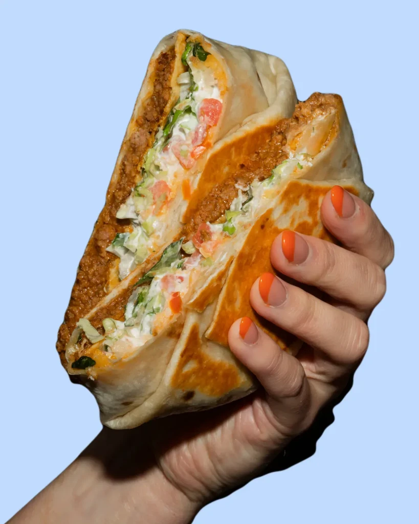 Hand holds cut burrito revealing layers of meat, sauce, lettuce, tomatoes.