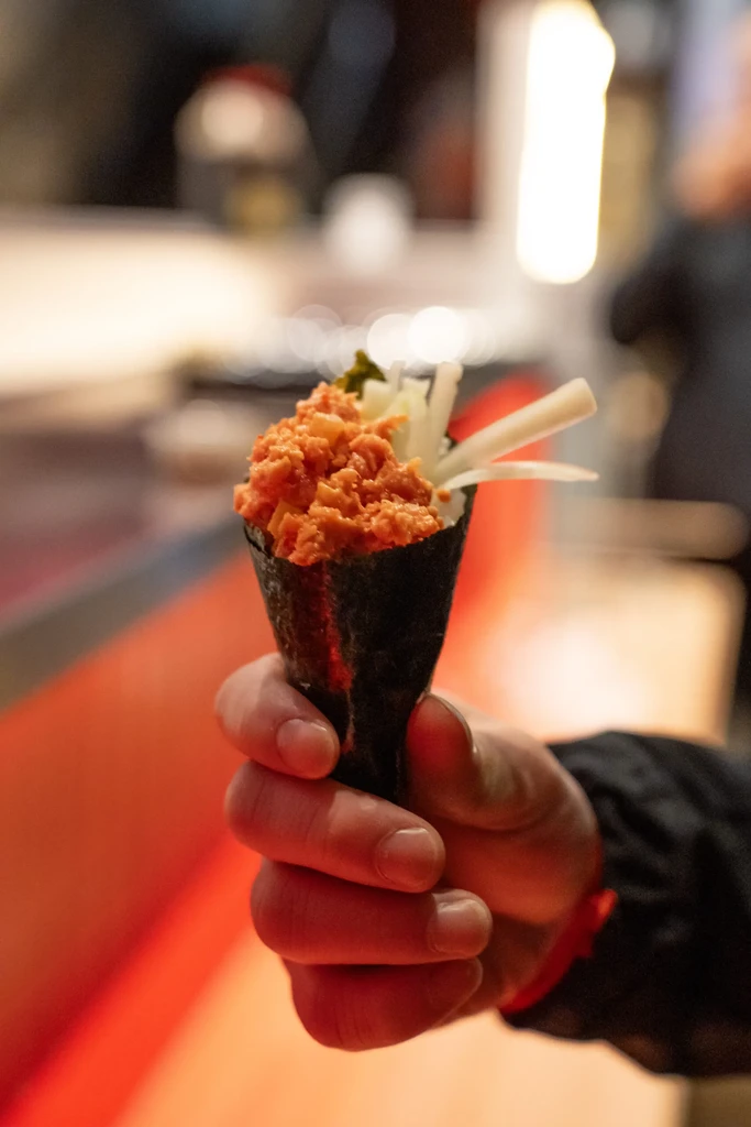Hand holding a temaki sushi with orange-red spicy tuna filling.