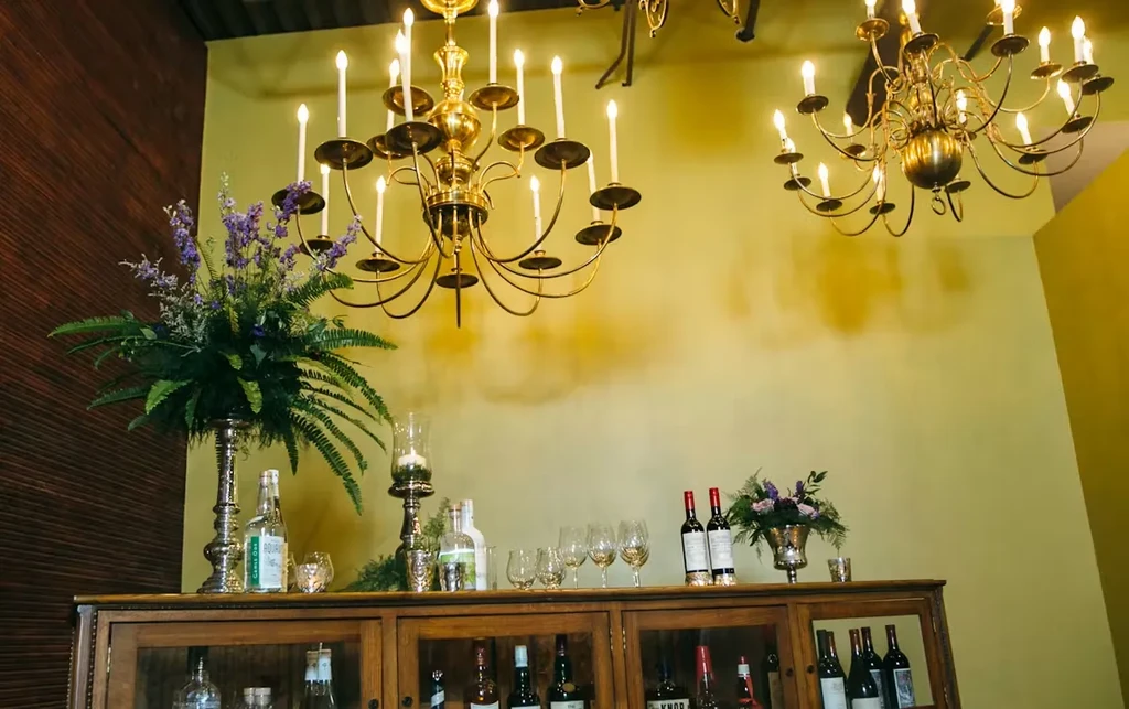 Ornate brass chandeliers illuminate a wooden bar with liquor and flowers.