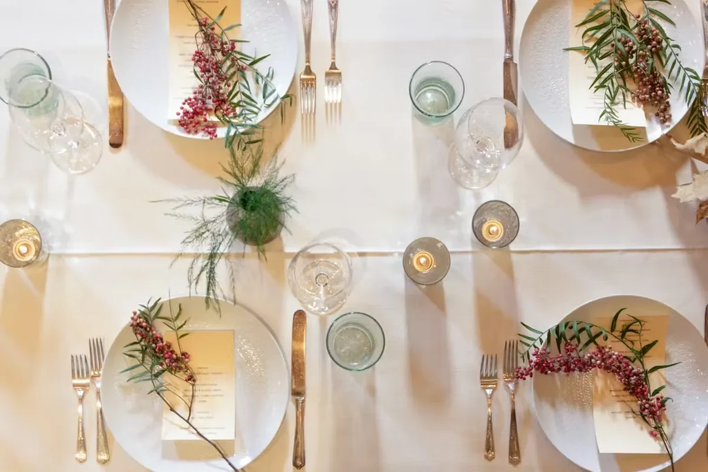 Dining table set for four with white plates, candles, and greenery.
