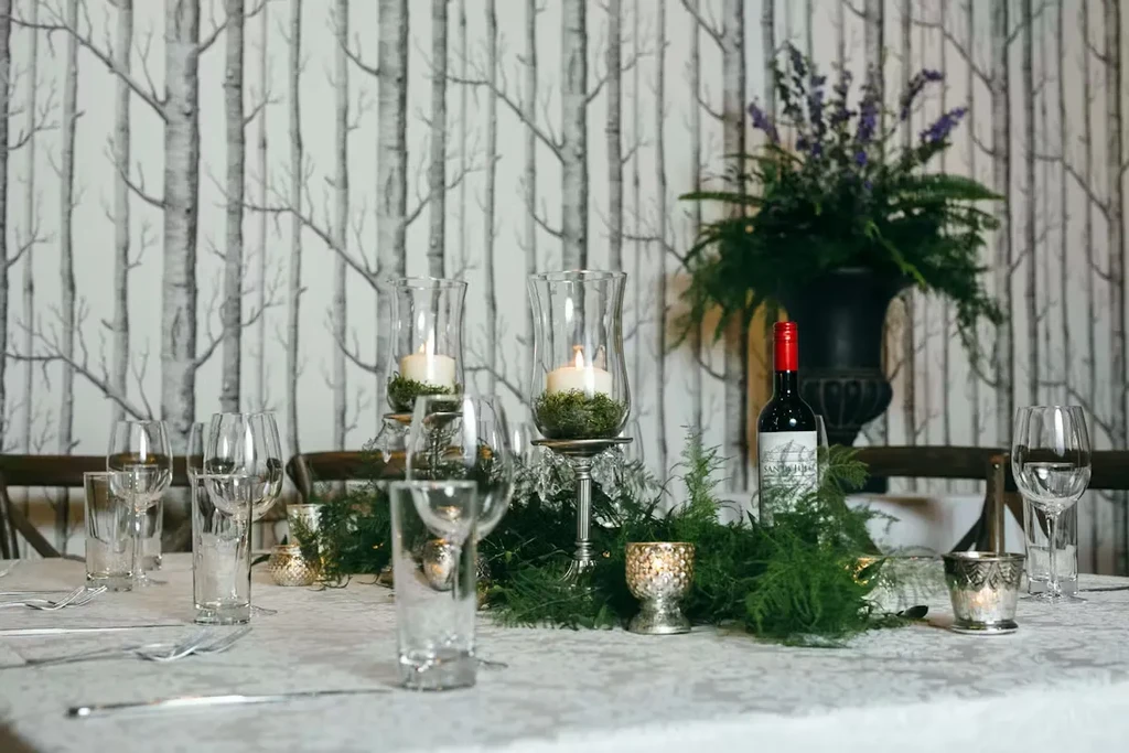 Elegant dining table with fern garland, lit candles, wine, and birch wallpaper.
