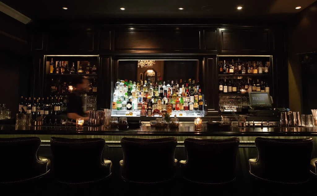 Dark, sophisticated bar interior with liquor display and blurred bartender.