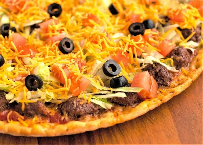 Taco pizza with meat, cheese, lettuce, tomatoes, and olives on wood.