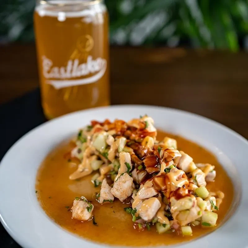 White plate of diced meat, greens, sauces, and Eastlake beer on wood.
