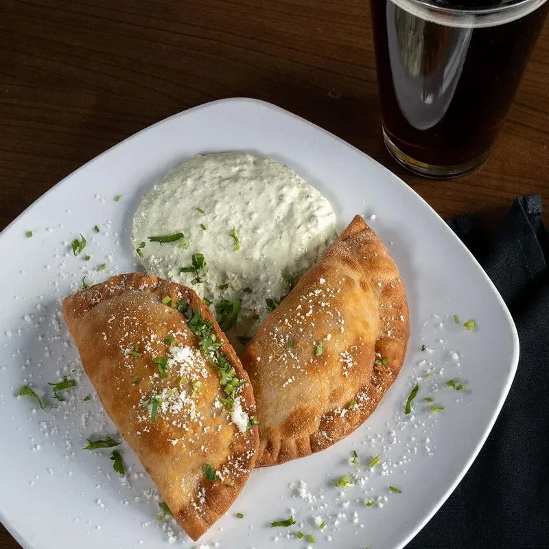 Two golden crescent pastries with cheese, herbs, dipping sauce, dark beverage.