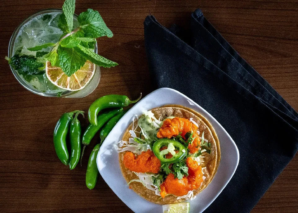 Overhead view of a taco, cocktail, and green chili peppers on wood.