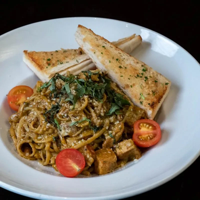 White plate holds pasta with creamy sauce, protein, tomatoes, and toast.