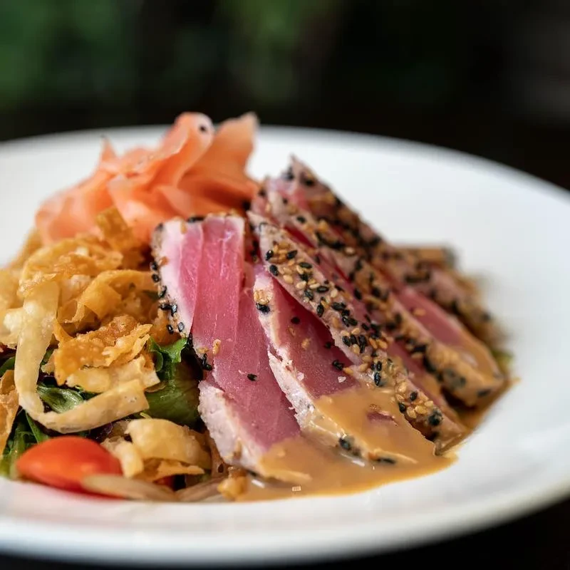 Seared tuna slices with sesame crust, creamy sauce, salad, and pickled ginger.