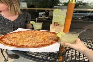 Round cheese pizza and margaritas on a black mesh patio table.