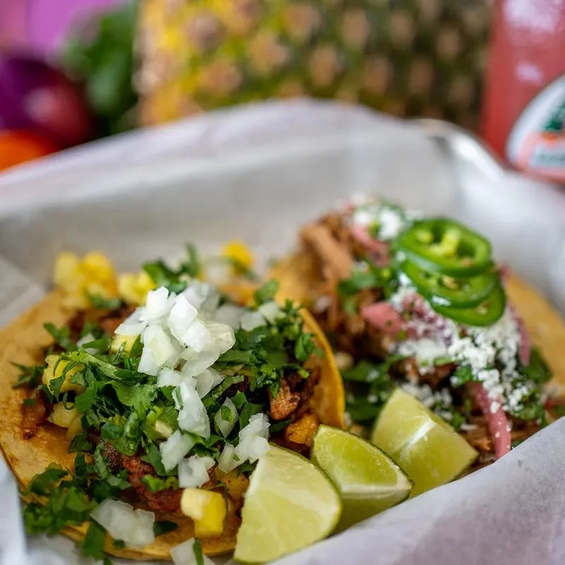 Two colorful tacos in a basket, garnished with lime, cilantro, and cheese.