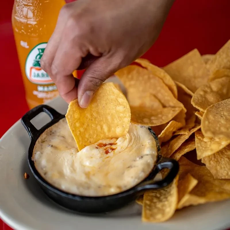 Hand dipping a tortilla chip into queso in a skillet with Jarritos.