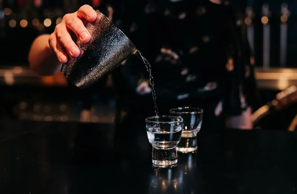 Hand pours clear liquid from jigger into glass in dim bar.