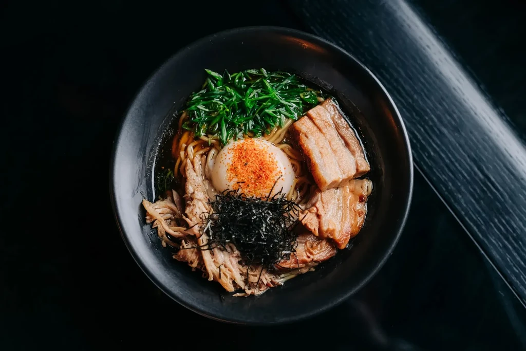Overhead: dark ramen bowl with pork belly, soft egg, scallions, and nori.