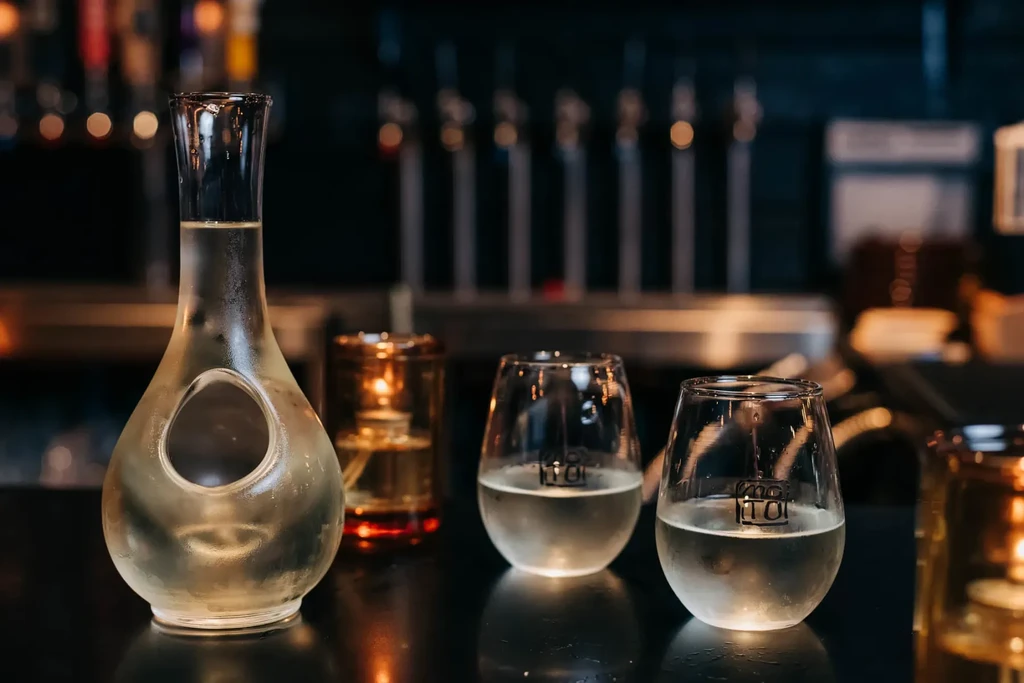Frosted decanter, two glasses with clear liquid on a dark bar.