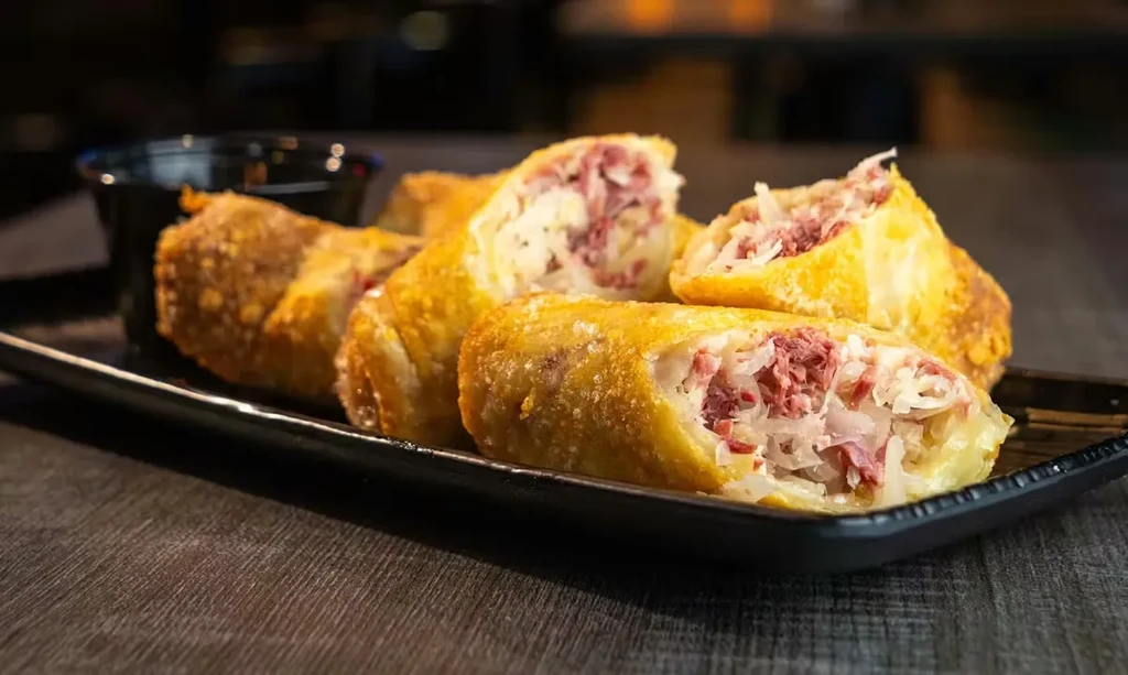 Deep-fried corned beef and cabbage rolls with sauce on black plate.
