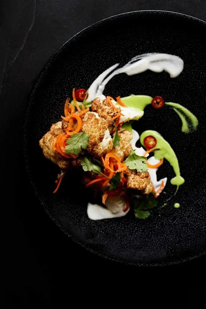 Golden-brown roasted cauliflower florets, carrots, cilantro, two sauces, black plate.