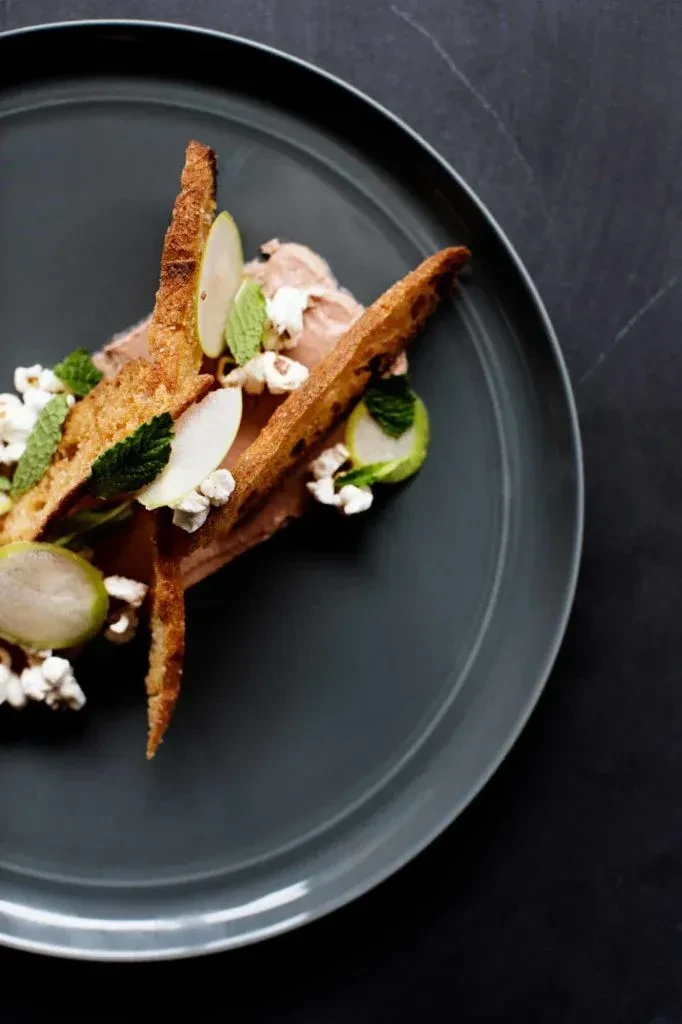 Elegantly plated creamy dish with crispy toast, green fruit, mint, and grains.