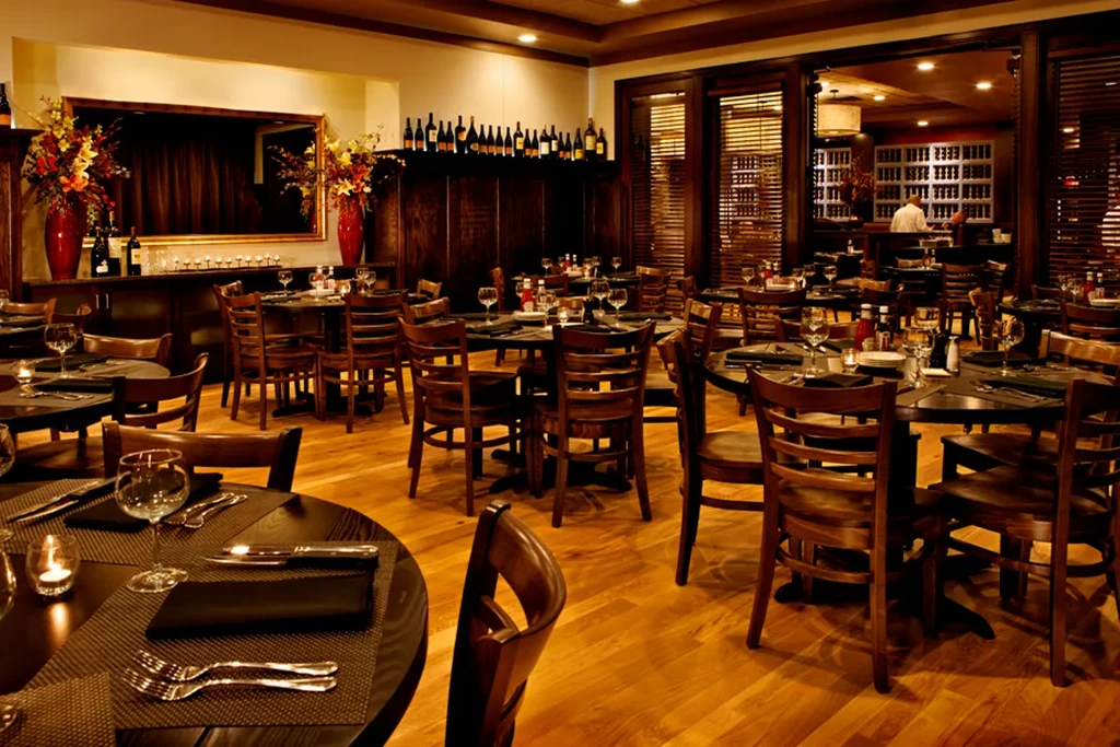 Upscale restaurant dining room with numerous set tables, warm lighting, wine bar.
