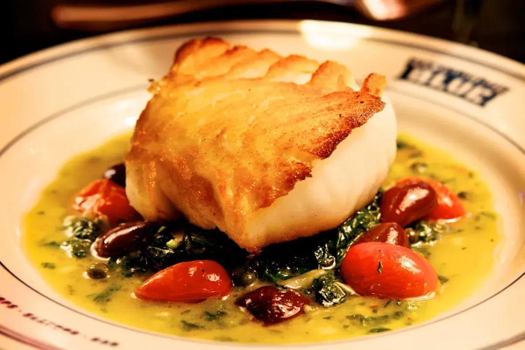 Plated white fish with golden crust, greens, tomatoes, olives, and sauce.