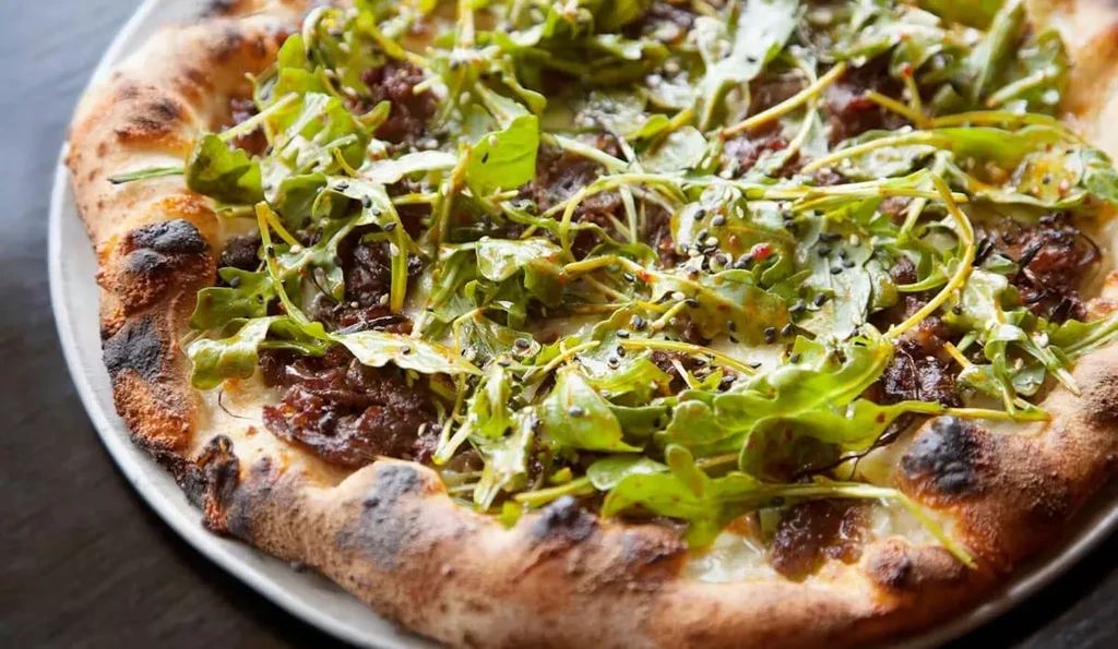Gourmet pizza with charred crust, arugula, savory toppings, and sesame seeds.
