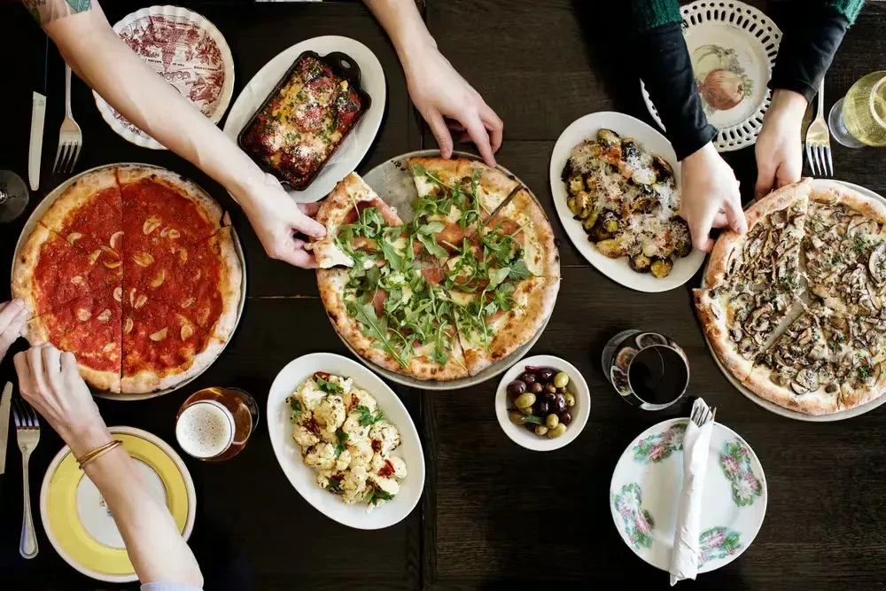 People share pizzas, pasta, roasted vegetables, and drinks at a table.