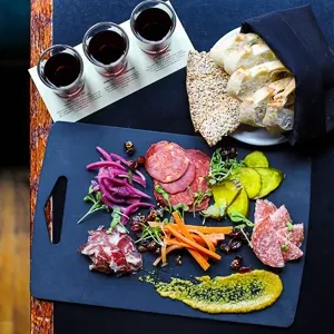 Top-down charcuterie board with meats, vegetables, wine, bread on a dark table.