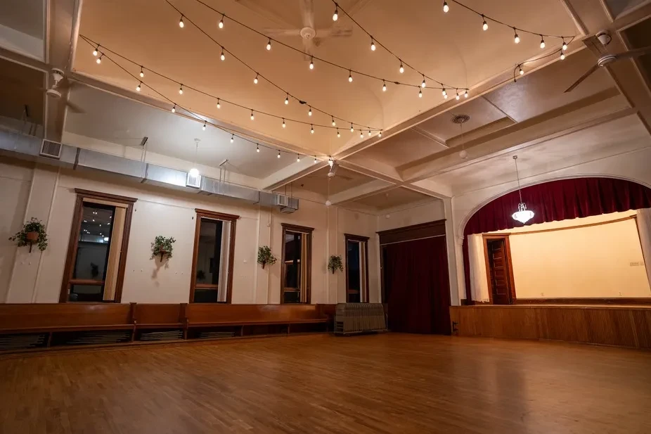 Warm-toned empty event hall with string lights, stage, and hardwood floor.