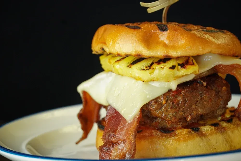 Close-up gourmet burger with beef, bacon, melted cheese, and grilled pineapple.