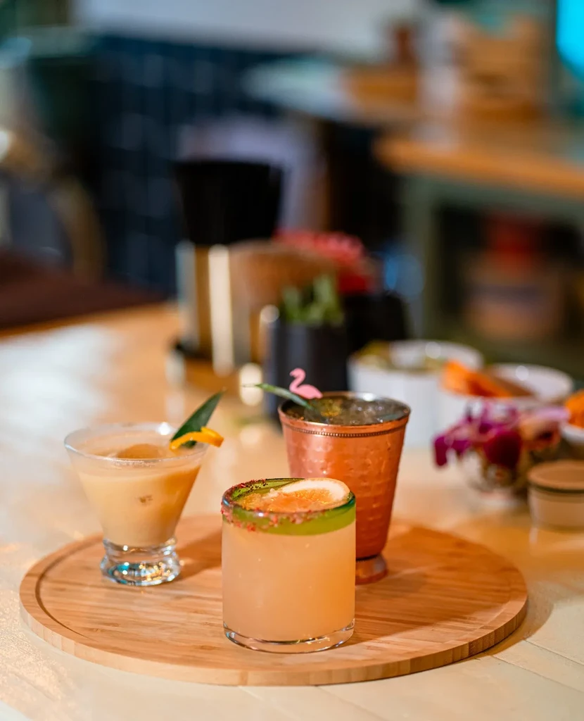 Three garnished cocktails on a wooden tray in a bar setting.