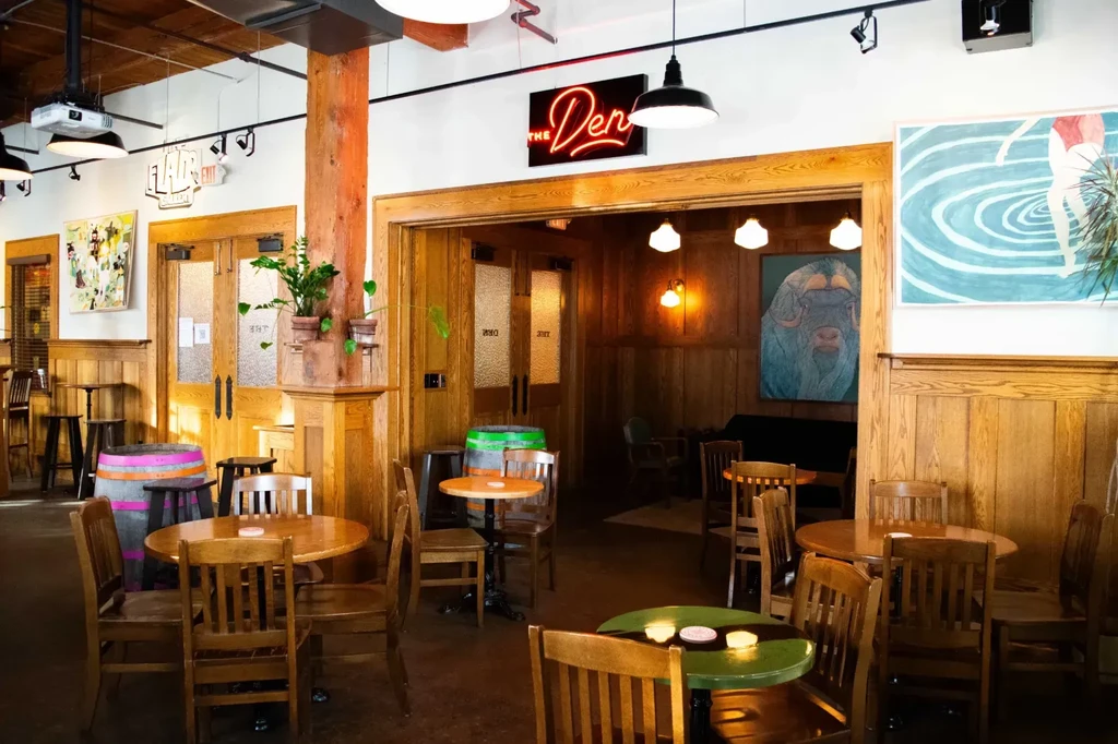 Rustic-modern restaurant interior with wood, barrels, and 'THE Den' neon.