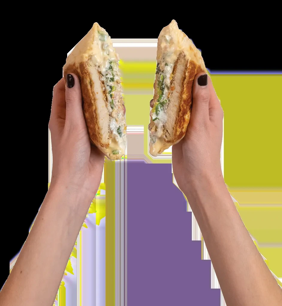Hands hold a large, triangular chicken flatbread sandwich with green filling.