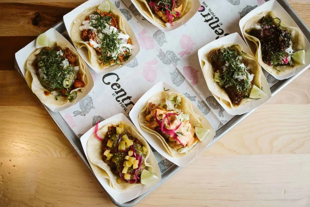 Overhead view of eight distinct tacos in paper boats on a tray.