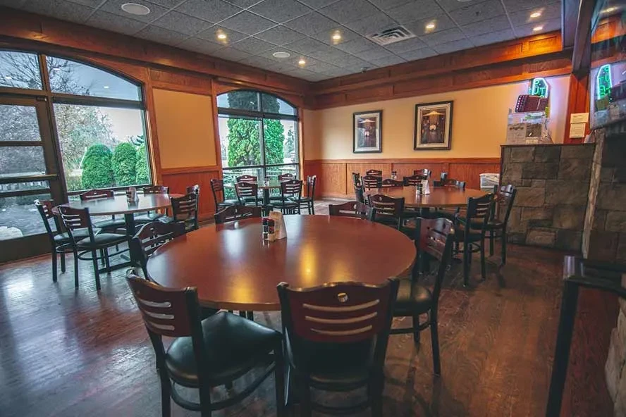 Empty restaurant with wooden tables, arched windows, and outdoor greenery.