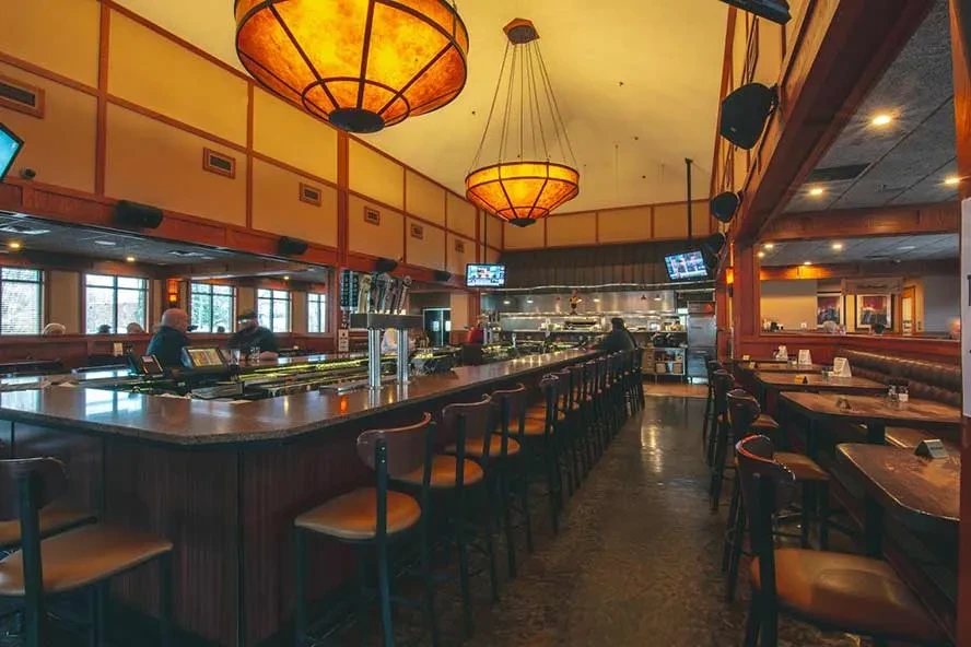 Warmly lit casual restaurant interior with long bar, booths, and open kitchen.