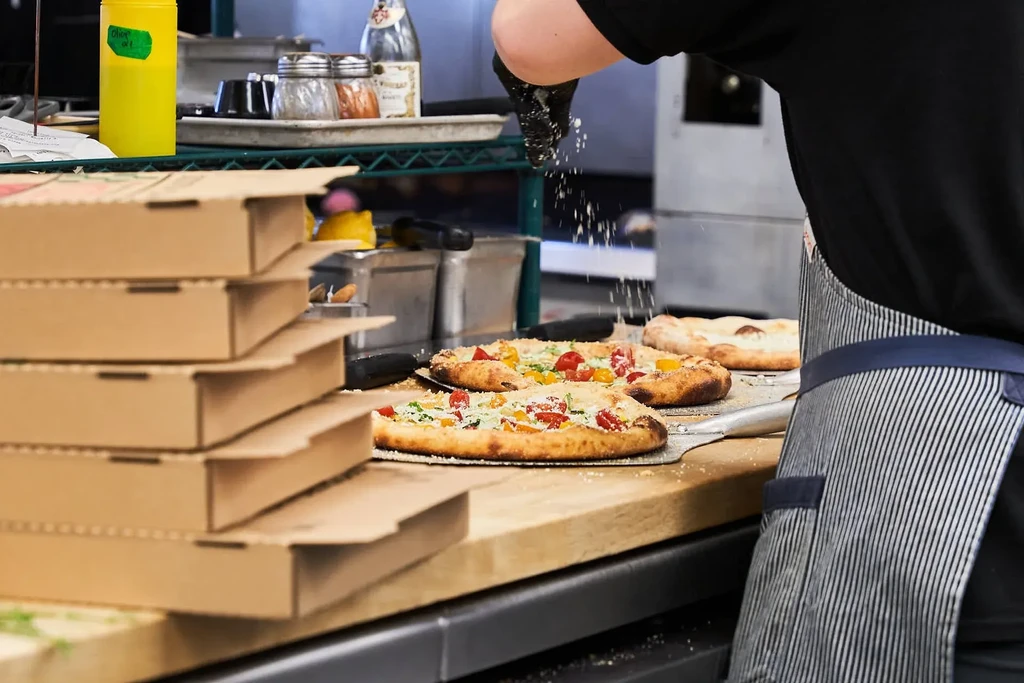 Chef sprinkles toppings on three fresh pizzas in a bustling kitchen.
