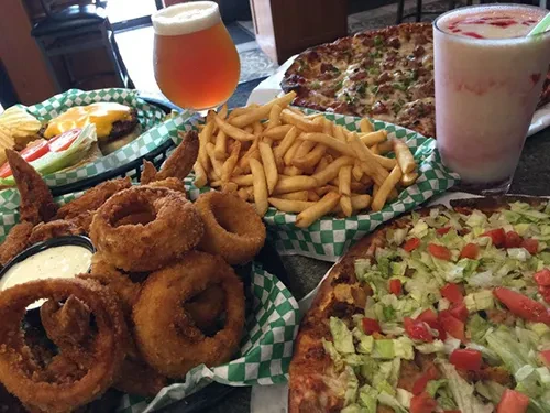 Table spread of cheeseburger, fries, wings, two pizzas, beer, and milkshake.