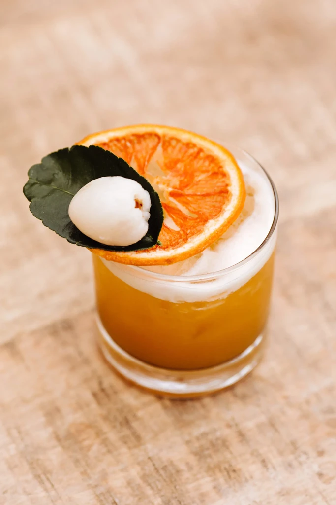 Golden-yellow cocktail, frothy, with citrus, leaf, and lychee garnishes.
