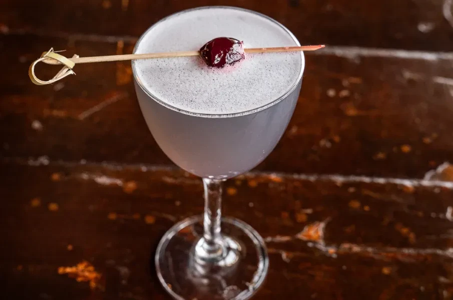 Light purple cocktail in coupe glass with foam and cherry garnish.