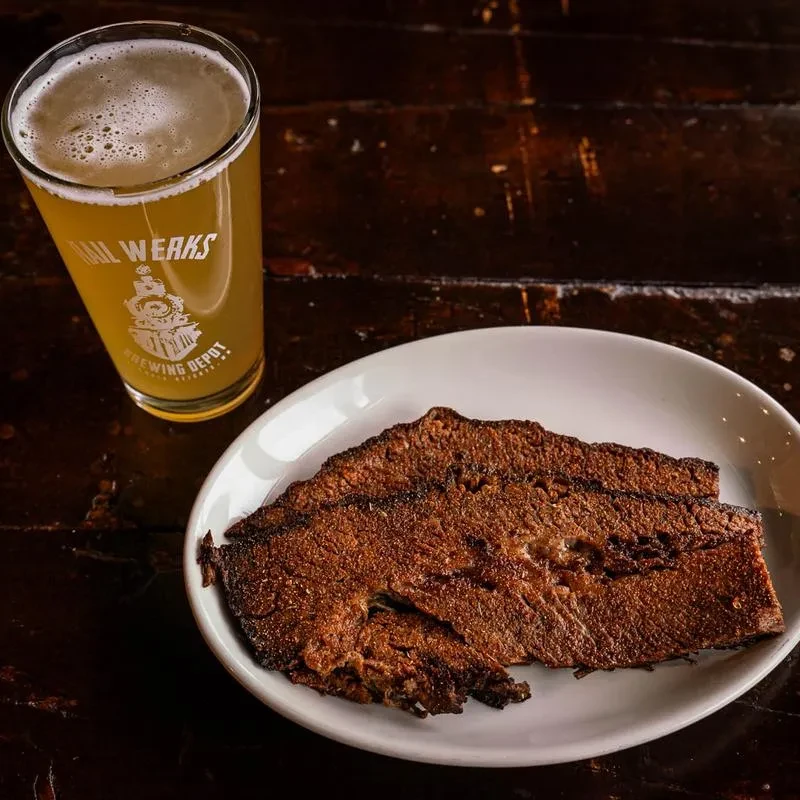 Rail Works beer and sliced dark-crusted meat on a rustic wooden table.
