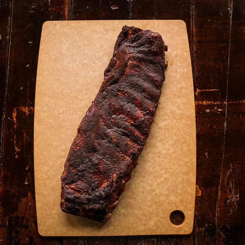 Dark, bark-covered smoked ribs on a light cutting board, rustic table.