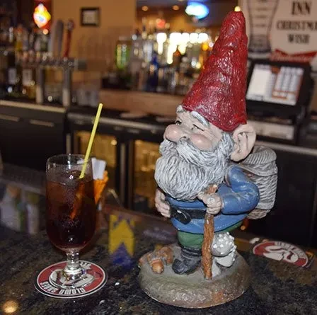 Detailed gnome statue and iced drink on a dim bar counter.