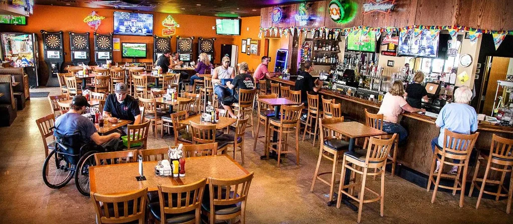 Lively sports bar interior featuring patrons, TVs, dartboards, and a bar.