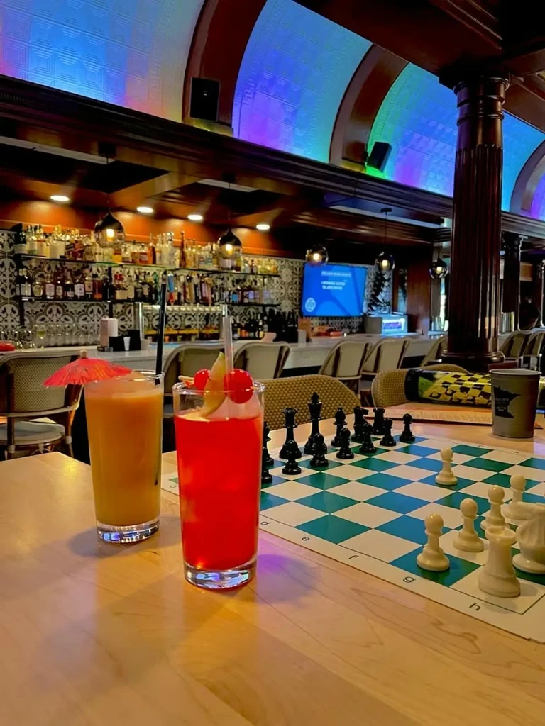 Table with two colorful drinks and a chessboard in a vibrant bar.