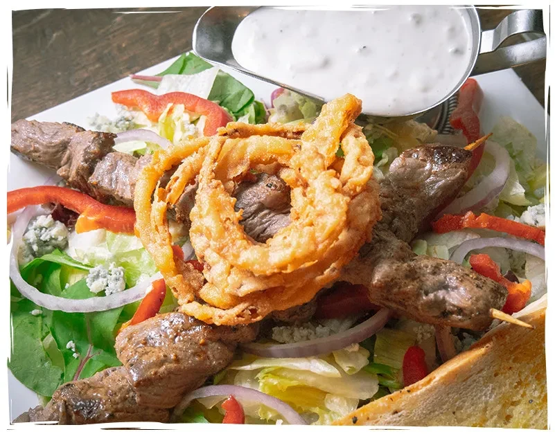 Hearty salad topped with beef skewers, crispy onion rings, and dressing.