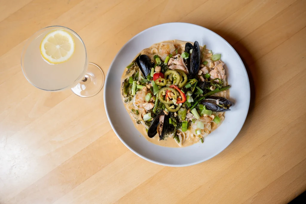 Overhead: creamy seafood noodles, vegetables, and lemon cocktail on wood.