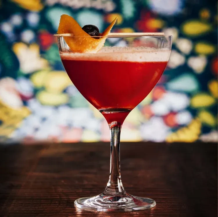 Red cocktail in coupe glass, with foam, twisted orange peel, and berry.