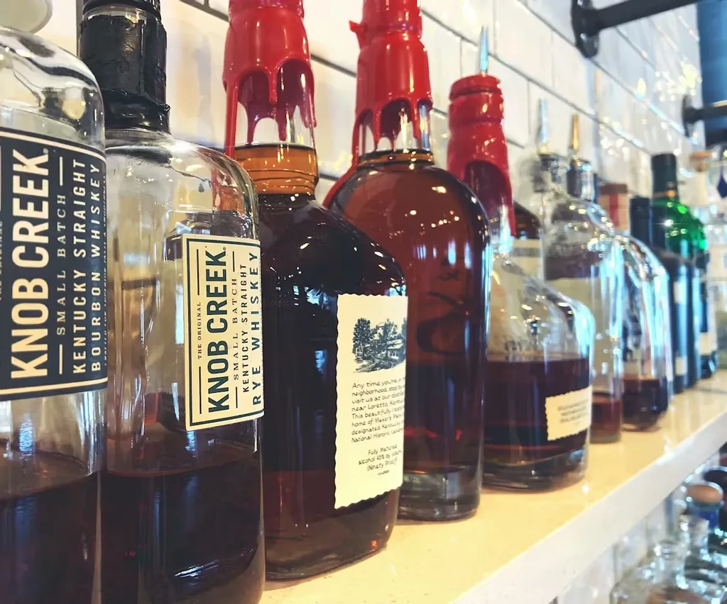 Bourbon and whiskey bottles, including Knob Creek, with red seals on a shelf.